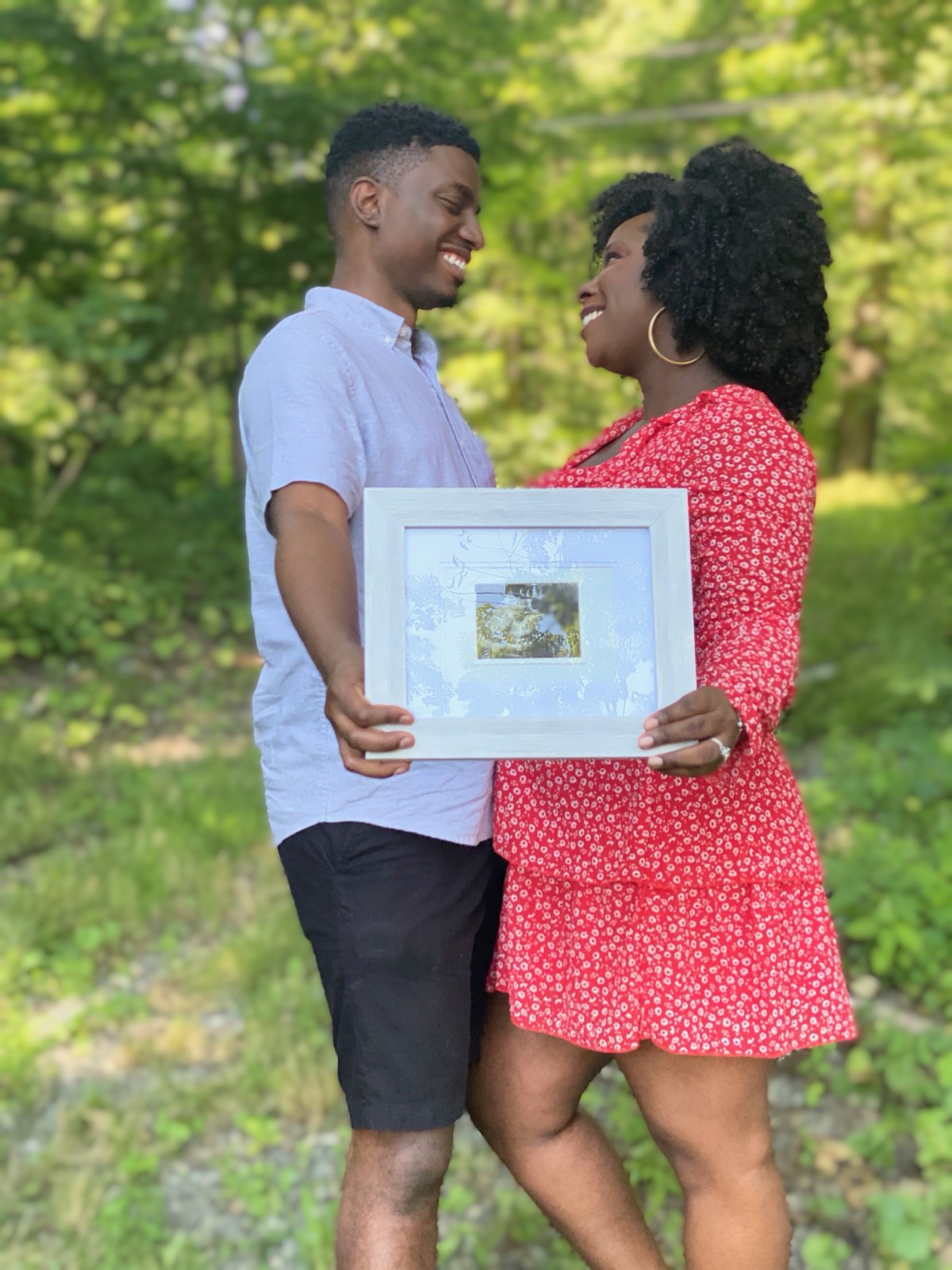An expecting couple smiling at each other outdoors while holding a framed sonogram.
