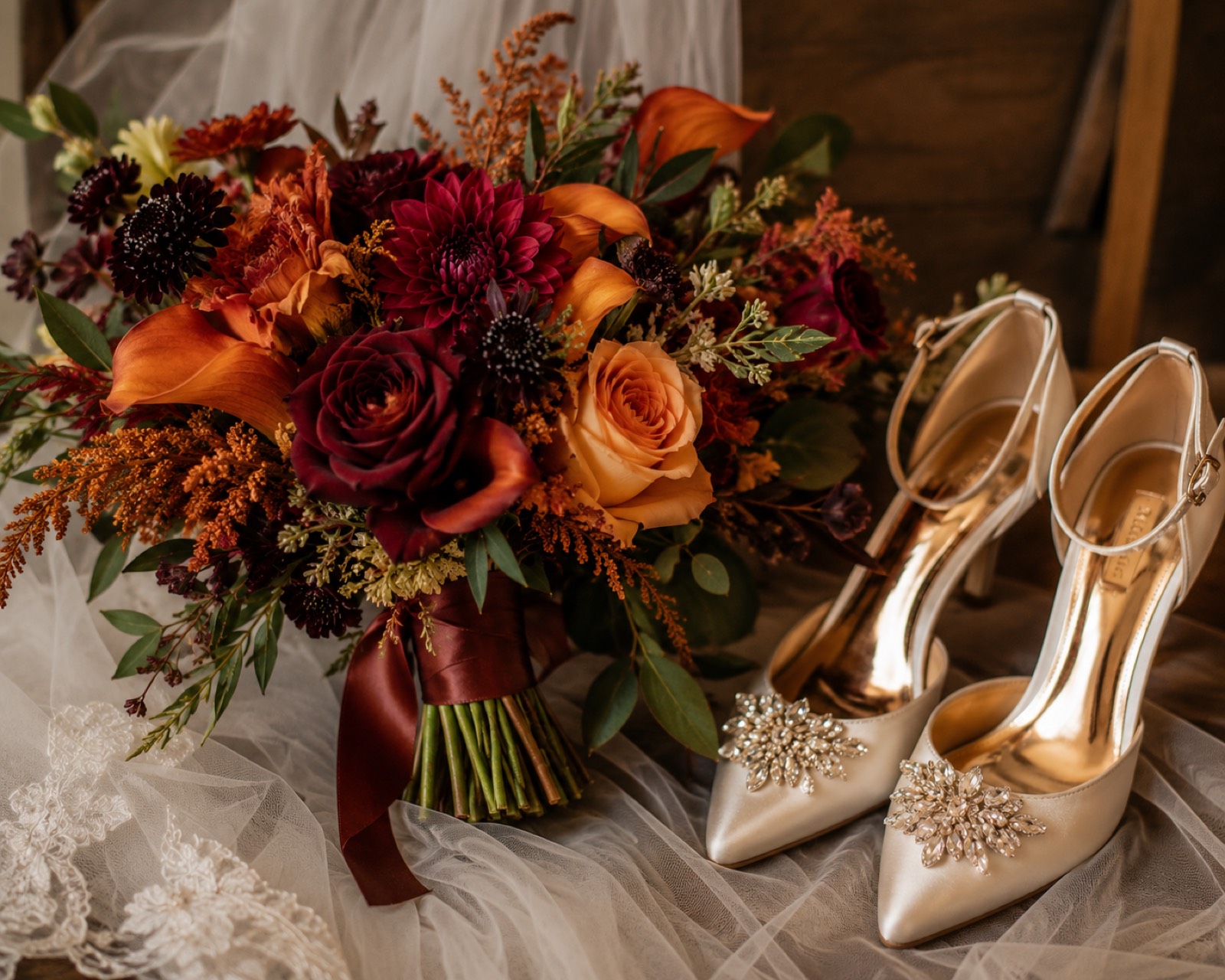 An autumn bridal bouquet of burgundy and orange blooms beside crystal-embellished ivory heels on tulle.
