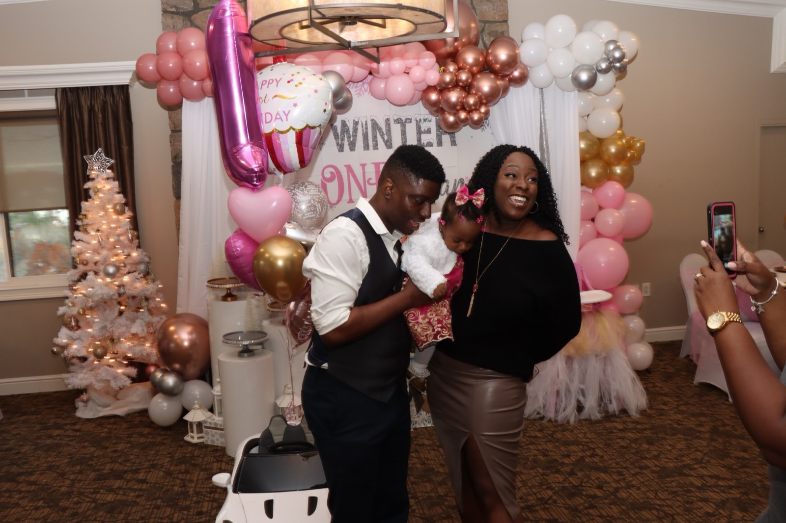 Parents holding their one-year-old in front of a pink-and-gold 'Winter ONEderland' balloon backdrop.