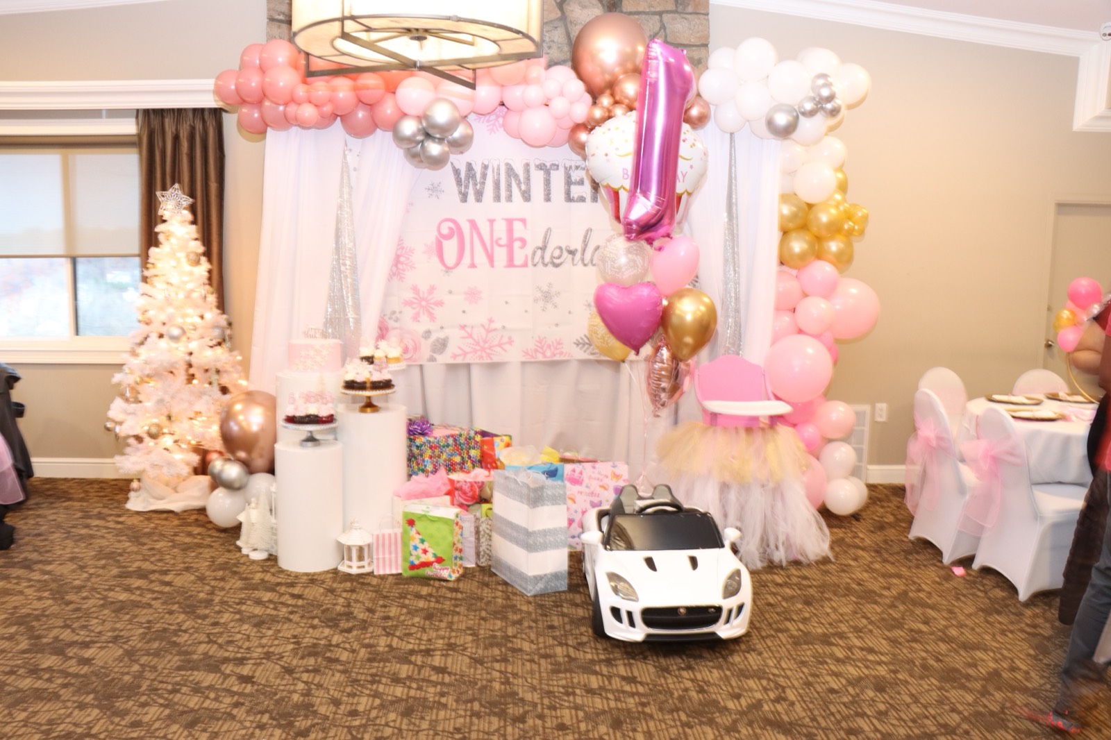 A 'Winter ONEderland' setup with a pink balloon arch, dessert table, gifts, and a child's ride-on car.