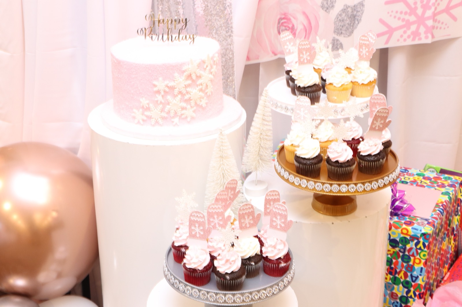 A pink snowflake birthday cake and mitten-topped cupcakes on crystal stands.