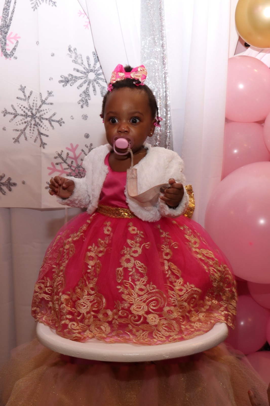 The one-year-old guest of honor in a pink-and-gold lace dress seated before a silver snowflake backdrop.
