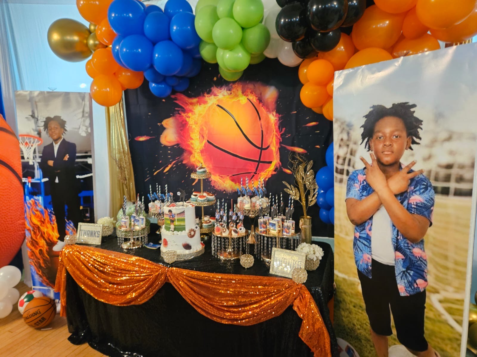 A basketball-themed dessert table with a flaming-basketball backdrop, balloon garland, and a sports-themed cake.